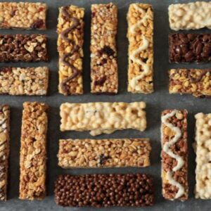 An assortment of 16 granola bars arranged in rows on a gray background. The bars vary in colors and textures, featuring chocolate drizzles, nuts, and crisped rice.